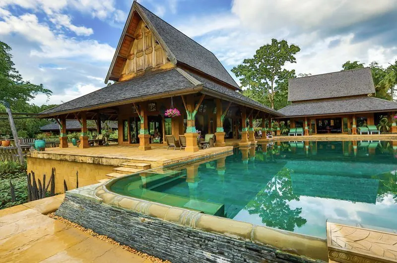 A Lanna-style teak pool villa with steep roofs beside a turquoise pool